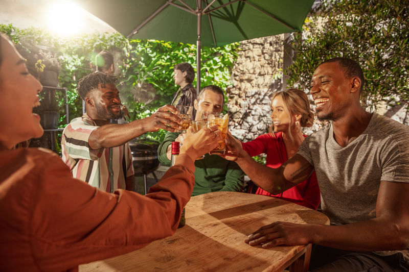 Group of friends sitting outdoors at a wooden table, smiling and raising whiskey cocktails in a celebratory toast under a patio umbrella