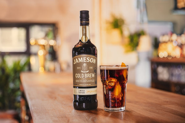 A bottle of Jameson Cold Brew Irish whiskey sits on a wooden bar beside a glass filled with iced cold brew whiskey cocktail, garnished with an orange twist, in a warmly lit bar setting.