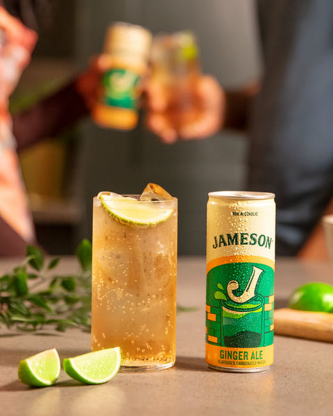 A tall glass of Jameson and ginger ale garnished with lime sits beside a can of Jameson Ginger Ale on a countertop, with lime wedges and two people toasting drinks in the background.