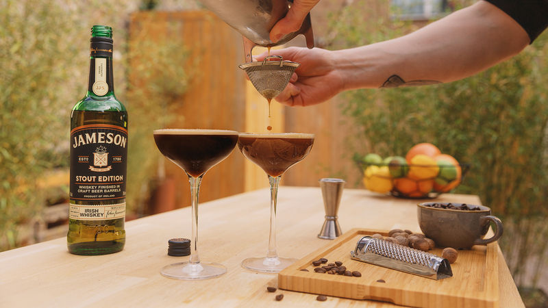 A bartender straining a rich Irish Espresso Martini into a glass next to a bottle of Jameson Stout Edition and coffee beans, illustrating a sophisticated spirit-forward digestif.