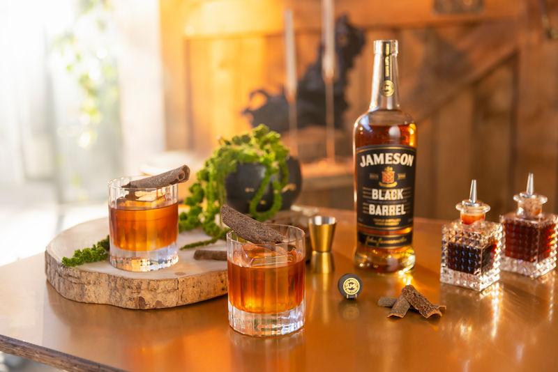 A bottle of Jameson Black Barrel whiskey standing next to two Old Fashioned cocktails served in glass tumblers with large ice cubes and cinnamon garnishes, alongside vintage bitters bottles.