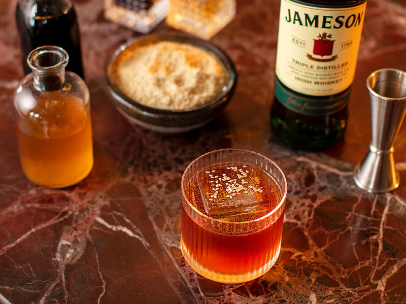 A close-up of a Cheung Fun Old Fashioned cocktail garnished with sesame seeds, sitting on a marble bar top next to a bottle of Jameson Irish Whiskey and a silver jigger.