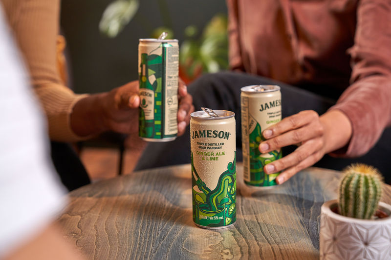 Close-up of friends holding cans of Jameson Ginger Ale and Lime Ready-to-Drink cocktails while socializing around a table.