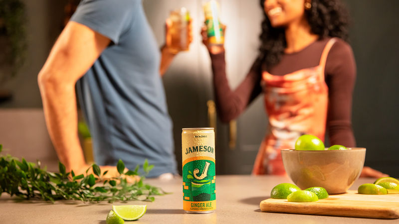 A Jameson Ginger Ale and Lime Ready-to-Drink can displayed on a kitchen counter next to fresh lime wedges, with a couple enjoying drinks in the blurred background.
