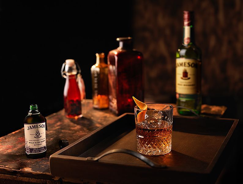 Whiskey cocktail with ice and orange peel on a tray, with Jameson bottle and bitters in the background