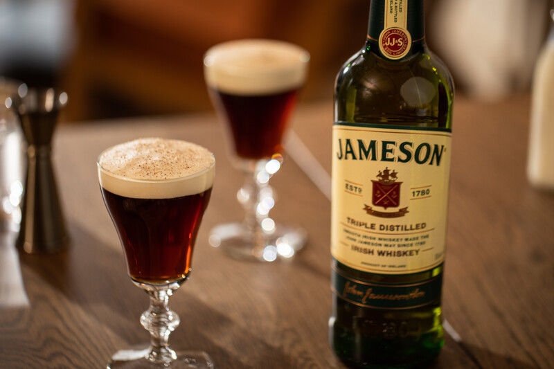 Two Irish coffee cocktails in stemmed glasses with a bottle of Jameson Irish whiskey on a wooden table