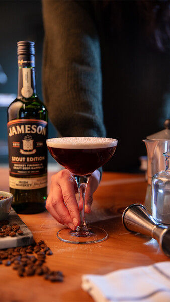 Bartender holding an Irish espresso martini made with Jameson Stout Edition, garnished with crema and surrounded by coffee beans.