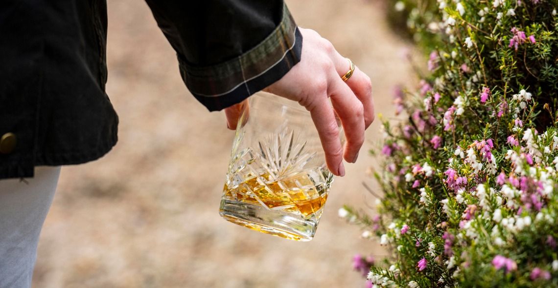 Close up of a hand carrying a whisky glass while walking in the grounds of Linnhouse surrounded by flowers.