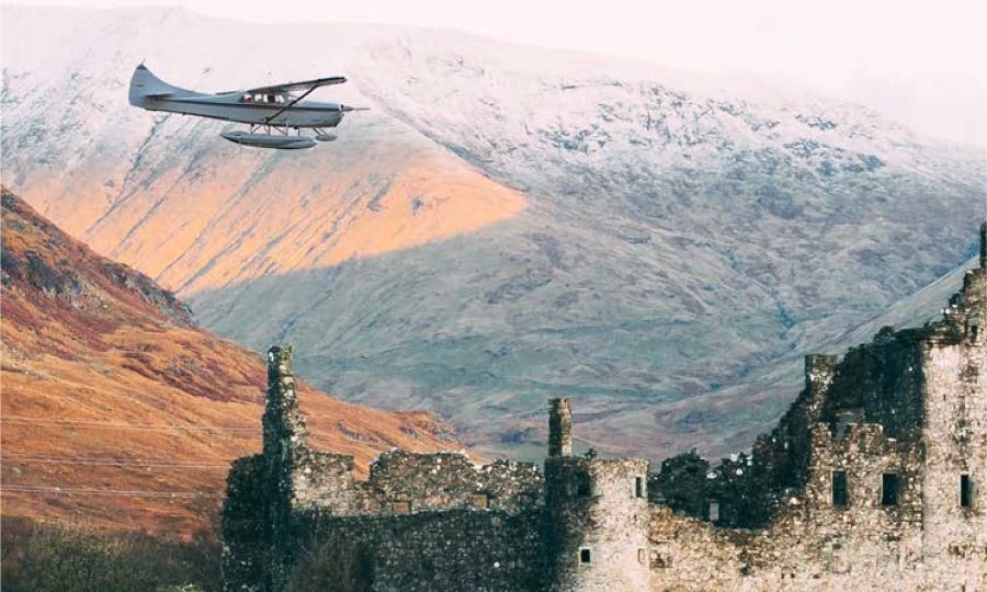 A small plane flying over a ruined Scottish castle, backed by mountains.
