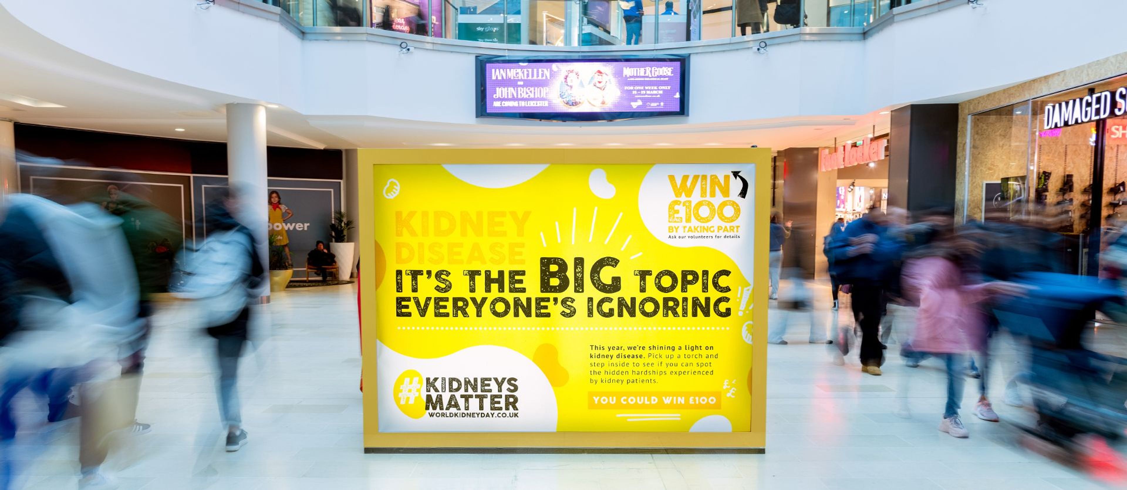 A large, illuminated stand for World Kidney Day in the middle of a shopping centre with people walking around it