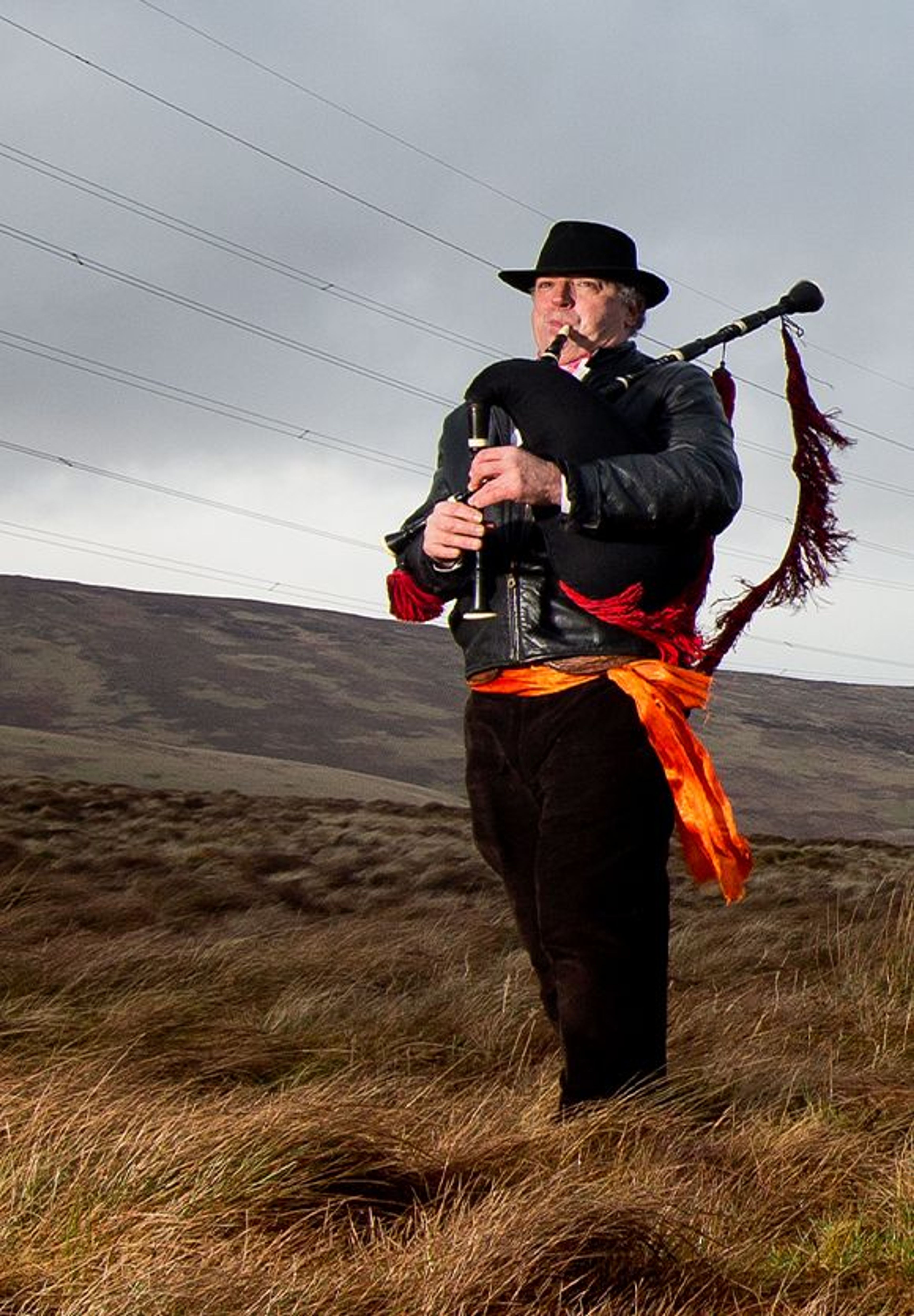 A man playing bagpipes stood in an expansive marsh with a white farmhouse in the background