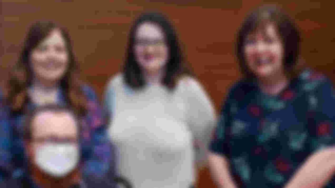 Three white adult women smiling and standing shoulder to shoulder plus one white man sitting in front wearing a medical face covering.