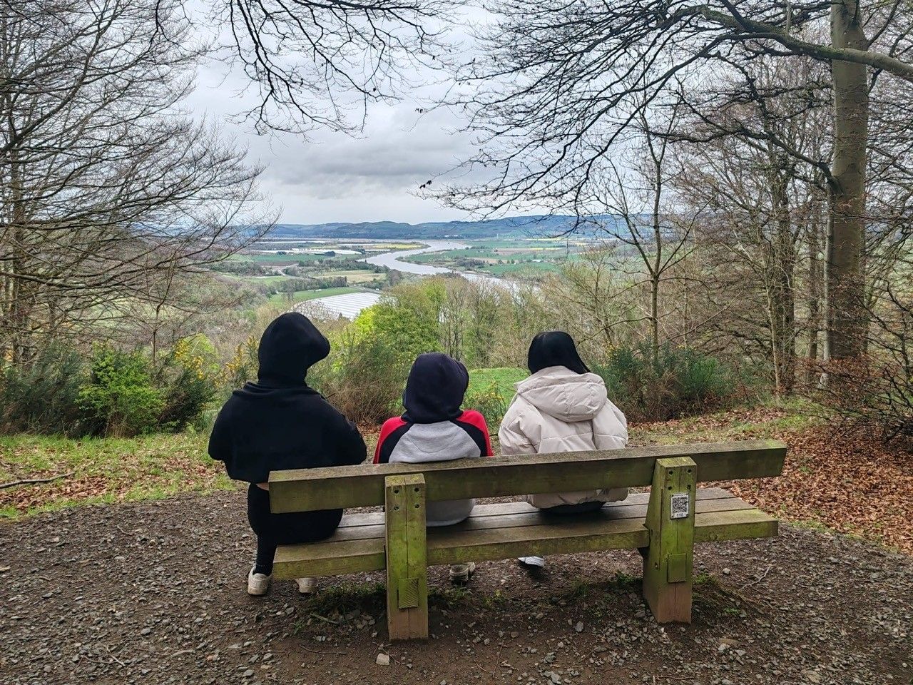 three people sitting on a bench with their backs to the camera looking at a beautiful countryside view