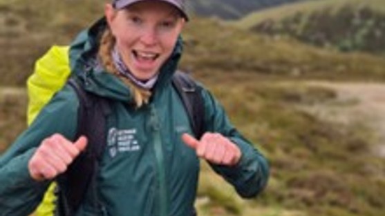 Young woman wearing green outdoor waterproof gear and a navy baseball cap smiling with two thums up. She is wering a backpack with a fluorescent bag cover and is standing in the hills of Perthshire.