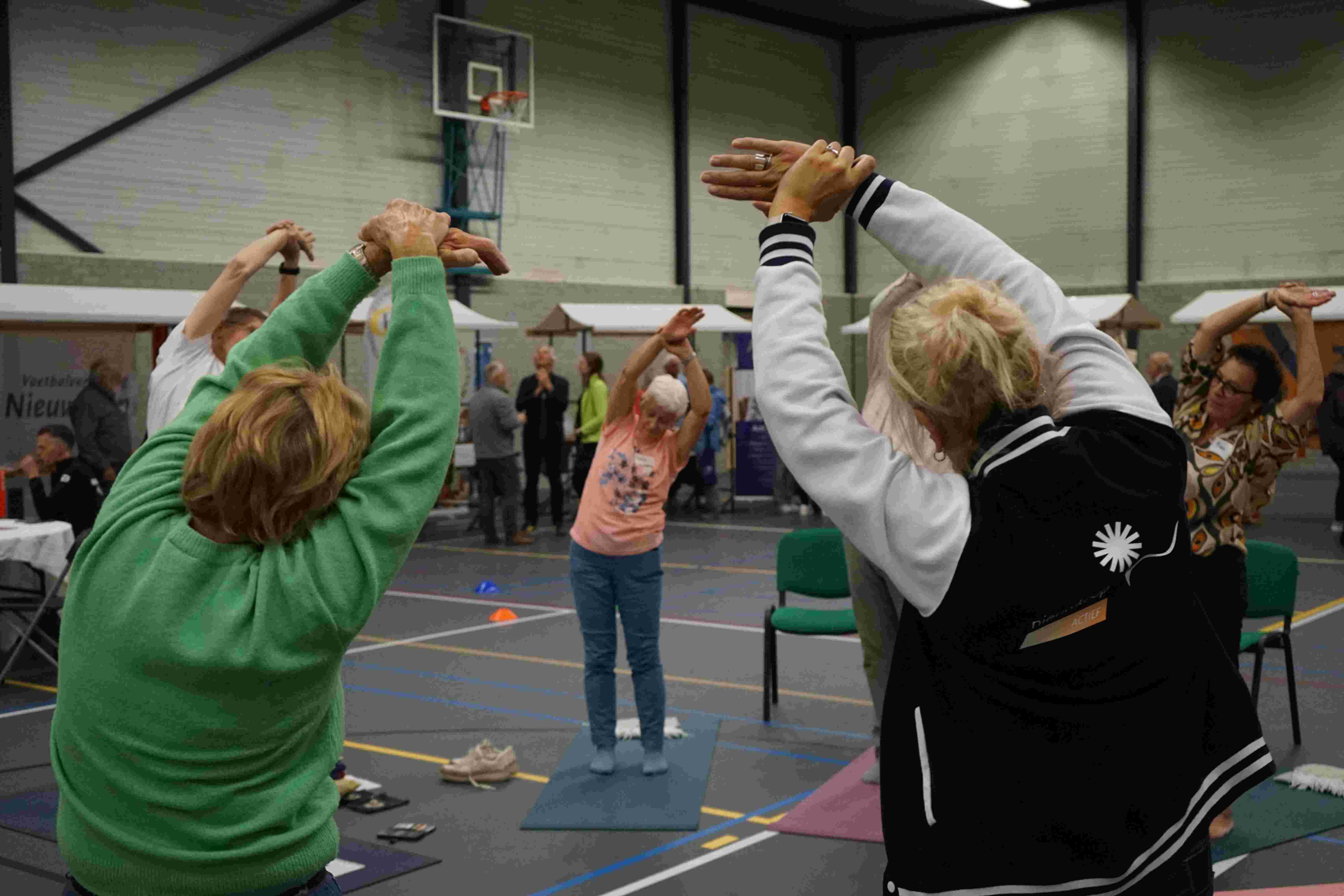Inwoners die meedoen met een demonstratie joga in een sporthal met kraampjes op de achtergrond