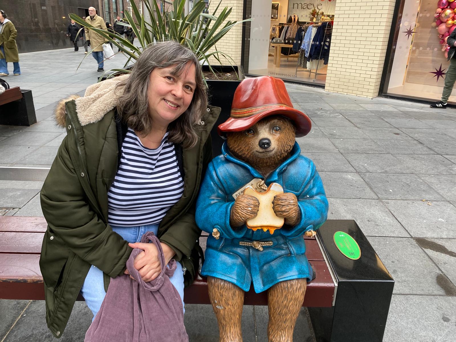 A photo of Christine sat next to a Paddington Bear statue. Chrstines has shoulder length brown hair and is wearing a stripey top and a green jacket.