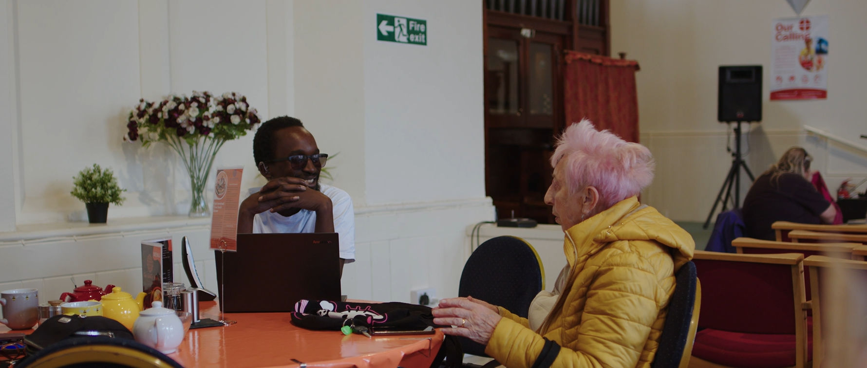 Photograph of Q, who features in the story sitting at a table talking to an older woman with pink hair.