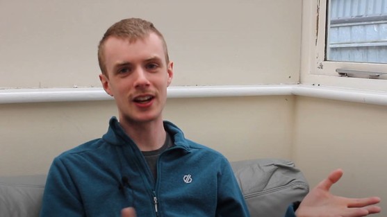 We see Alex, he is smiling and wearing a blue zip up top and has his hand up like he's in the middle of explaining something. He is sat down next to a window.