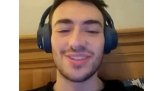 We see a young man smiling at the camera, he is wearing headphones and has a black t shirt.