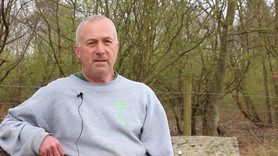 Alistair is sat on a bench in a wood, we can see trees behind him. He is wearing a grey sweatshirt and has short grey hair.