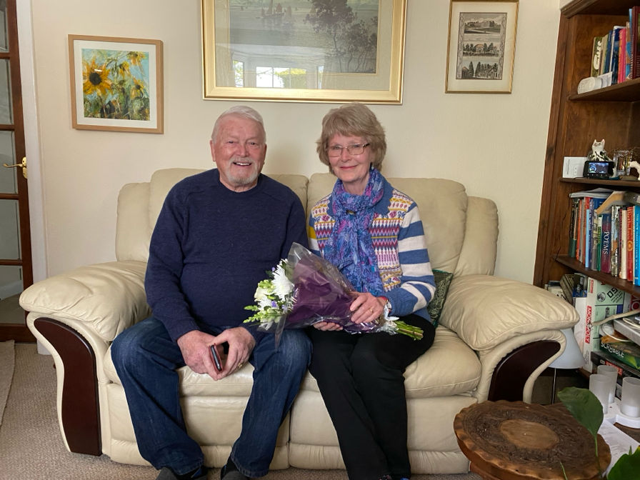 Anne and Bruno are sat on a cream couch in their lounge. They are both smiling to camera. Anne is wearing a stripy top, a blue scarf, black trousers and is holding a bunch of flowers. Bruno is wearing a navy jumper and jeans.