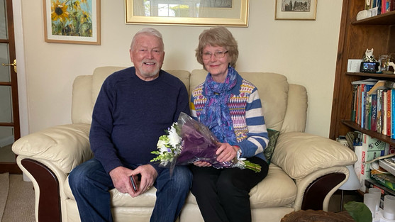 Anne and Bruno are sat on a cream couch in their lounge. They are both smiling to camera. Anne is wearing a stripy top, a blue scarf, black trousers and is holding a bunch of flowers. Bruno is wearing a navy jumper and jeans.