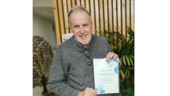 Duncan holding his volunteer award certificate. Duncan in smiling and wearing a grey jumper. He has grey hair and a short beard. We see a plant behind him.