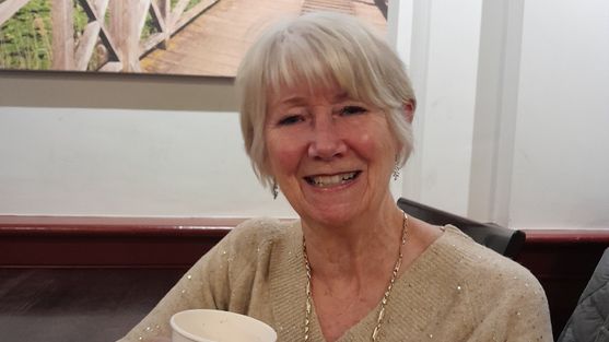 Beverly has short grey hair, she is smiling and is wearling a gold sparkly jumper. She is holding a cup of coffee.