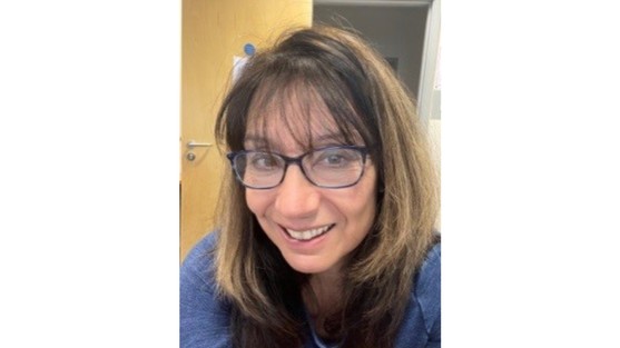 Kathy is smiling at the canera, she has long brown hair and is wearing glasses. She is wearing a blue jumper.