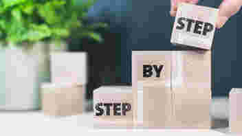 Wooden blocks on a table with a plant in the background. The words step by step can be seen on the blocks as they rise in a staircase fashion.
