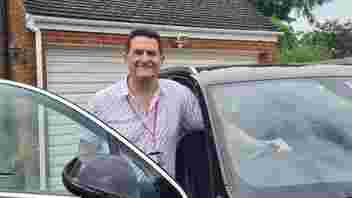 Man standing in a car door way, with one hand on wheel.