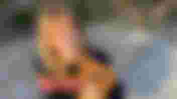 Woman with blond hair wearing blue jumper with stripes. She is smiling and kneeling down while holding a small dog