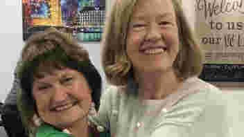 Image shows two volunteers dressed in Christmas jumpers hugging each other and smiling at the camera.