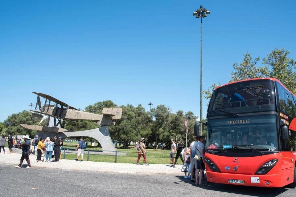  red double-decker hop-on hop-off sightseeing tour bus parked near a historic airplane monument and park area in Lisbon