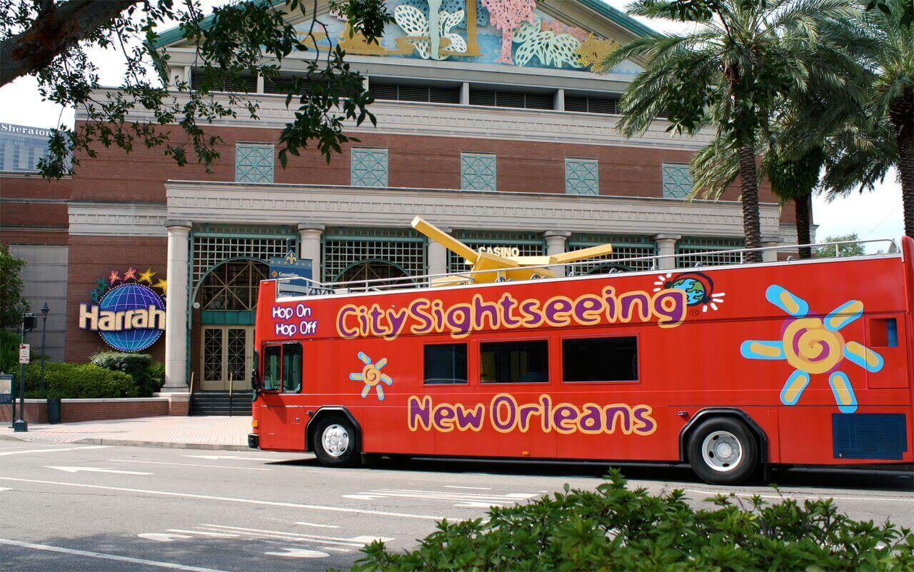 The image shows a bright red City Sightseeing hop-on, hop-off open-top double-decker bus driving past the Harrah's New Orleans casino building