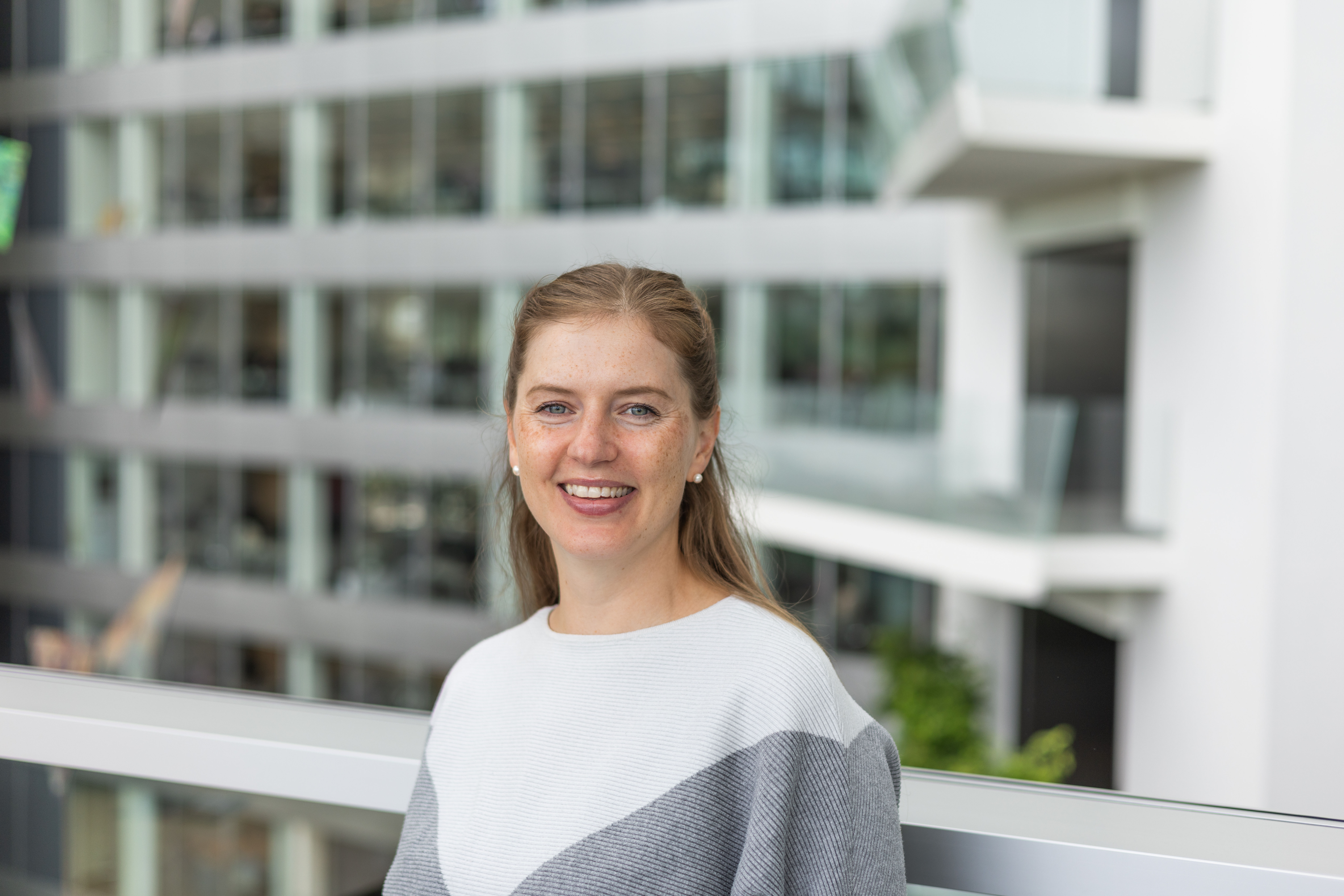 Smiling woman in a light gray sweater sits outdoors on a balcony with modern buildings in the background.