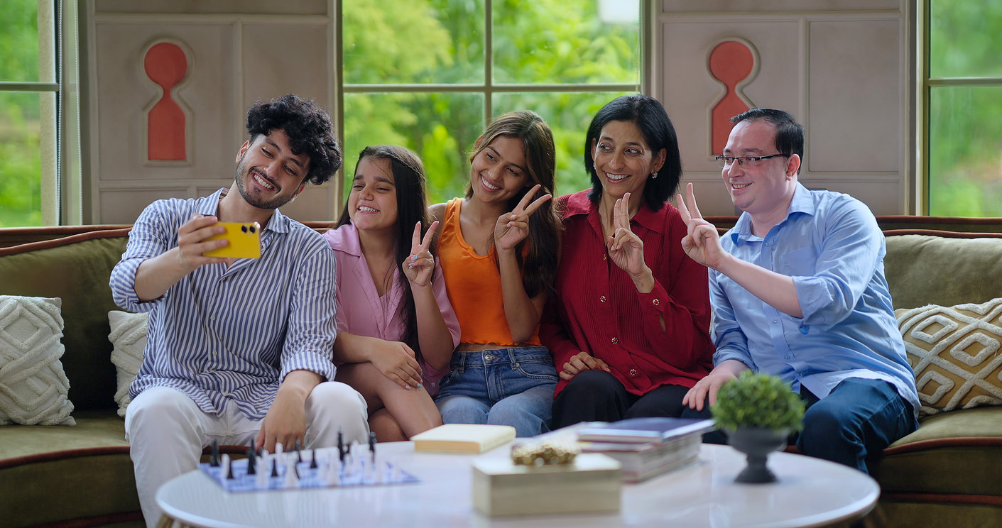 Indian happy mother father with young adult children sitting cozy sofa couch use mobile phone take selfie photo picture indoor house. Teen girl boy look cam make funny pose photo record video enjoy together