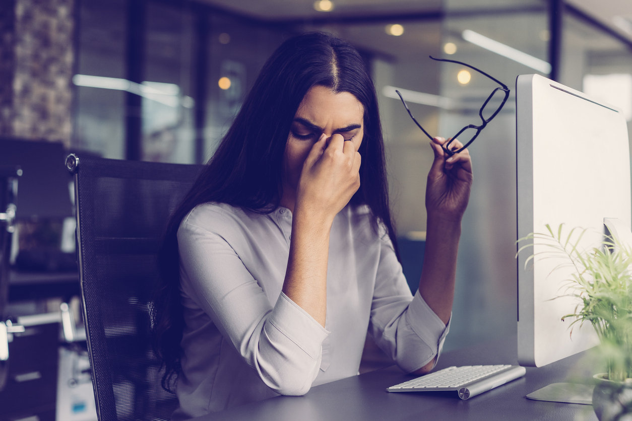 Portrait of depressed young Latin-American businesswoman sitting at computer in office holding glasses and rubbing eyes. Overworking concept