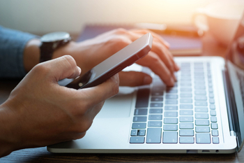 Man hand using mobile phone and typing keyboard of laptop computer on office desk workspace