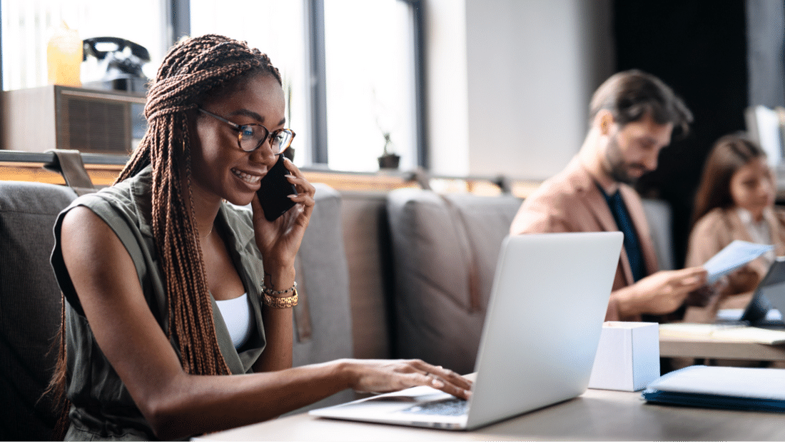 An African American woman working on her laptop while talking on the phone in a work space