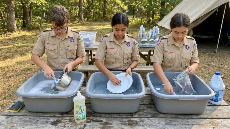 Scouts at a camp wash station using the three-basin system — one Scout scrubbing a pot in soapy water, another rinsing, and a third dipping utensils in a sanitize basin