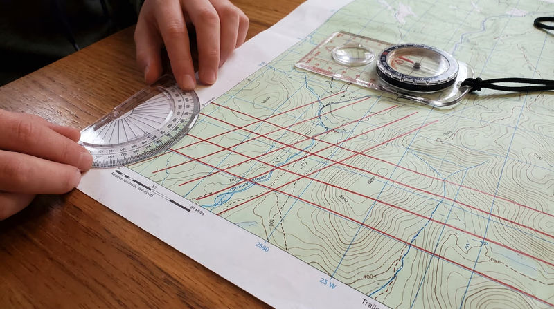 Hands drawing magnetic north-south lines on a USGS topographic map using a straightedge and red pencil, with a protractor at the starting point and several parallel red lines already drawn at a slight angle from the printed grid lines
