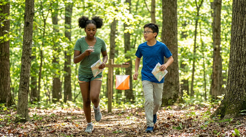 Two Scouts running through an open forest with orienteering maps in hand, one checking a compass while the other looks ahead toward a distant orange-and-white control marker flag