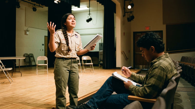 A teenage actor standing center stage during a rehearsal, script in hand, gesturing expressively while a director watches from the front row