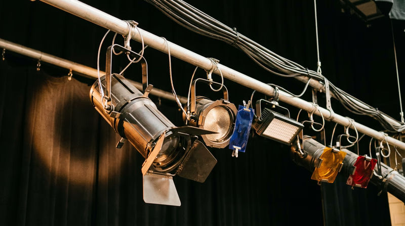 Various stage lighting instruments hanging from a lighting pipe above a stage, including an ellipsoidal, a fresnel, and LED fixtures, with colored gel frames visible