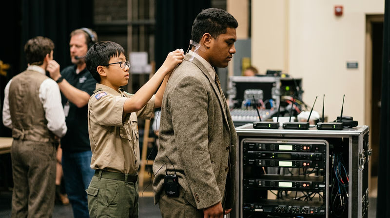 A sound technician backstage helping an actor adjust a wireless microphone pack attached to their costume, both looking focused