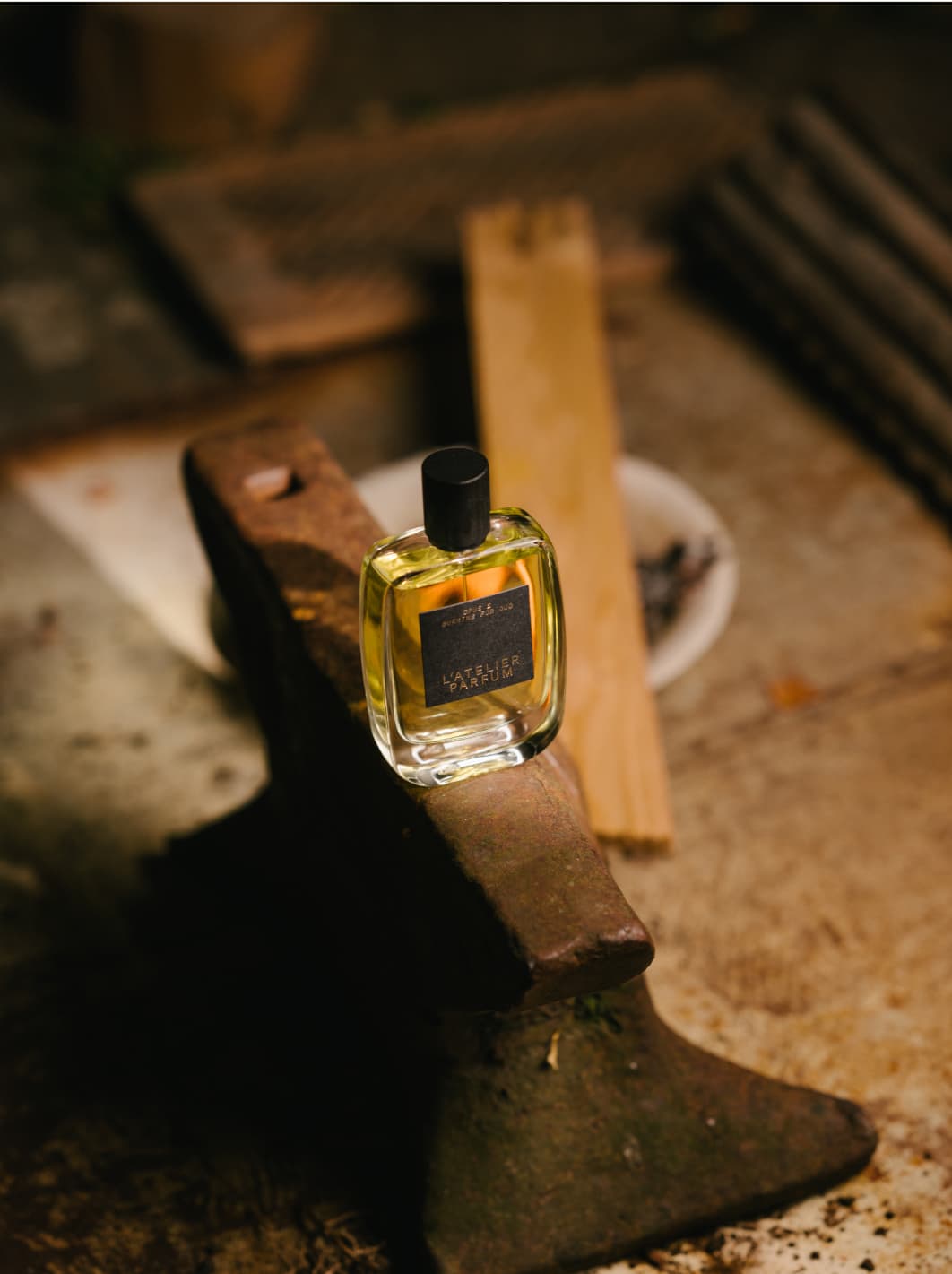 Flacon des Nischenduftes Borning for Oud von L'Atelier Parfum auf einem Amboss stehend mit brennendem Holz im Hintergrund