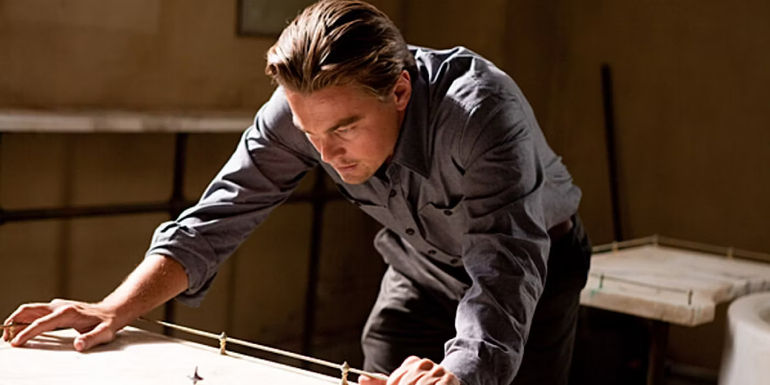 Cobb looks down at his spinning top with his hands braced on the table in Inception.