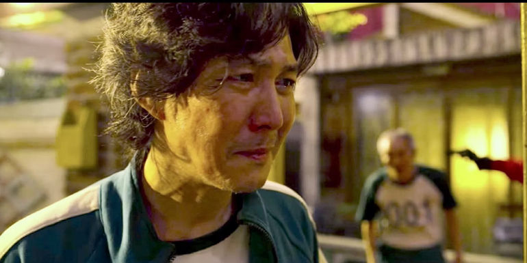 Gi-hun has tears in his eyes as II-Nam has a gun to his head behind him in Squid Games.