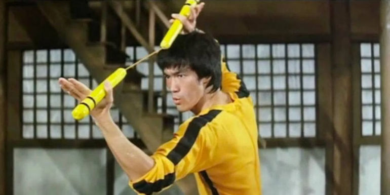 Bruce Lee as Billy Lo holding yellow nunchucks while wearing his signature yellow tracksuit in Game of Death's fight scene